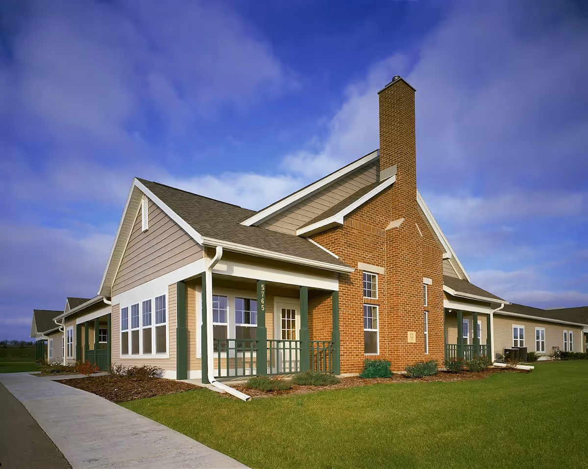 Exterior view of a single-story senior living facility building with beige siding, green trim, a brick chimney, and a covered porch. The building is surrounded by a well-maintained lawn and a sidewalk leading up to the entrance. The sky is partly cloudy with blue patches.