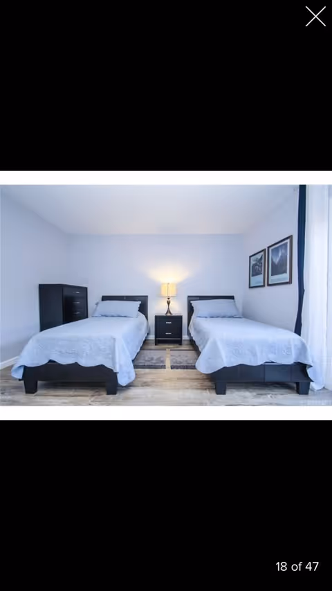 A bedroom with two single beds, each with light blue bedding and pillows. Between the beds is a small black nightstand with a lamp on it. To the left of the beds is a black chest of drawers. Two framed pictures hang on the wall above the bed on the right. The room has light-colored wooden flooring and light blue walls.