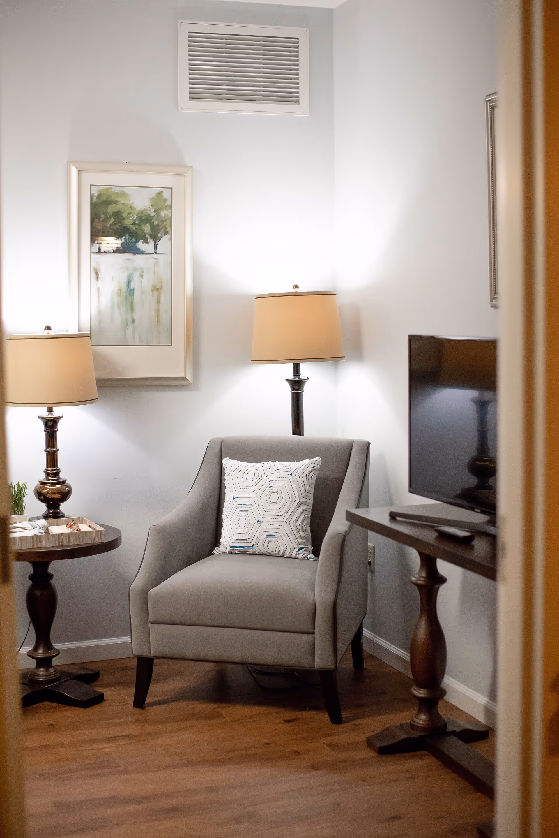 Gray upholstered armchair with a patterned pillow between two lamps and side tables, next to a TV and framed wall art.