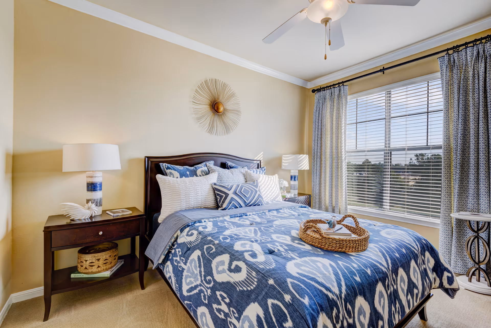 A cozy bedroom with a large window covered by patterned curtains. The bed has a dark wooden headboard and is made with blue and white patterned bedding and multiple pillows. There are two matching bedside tables with lamps and decorative items. A ceiling fan with a light hangs above the bed.