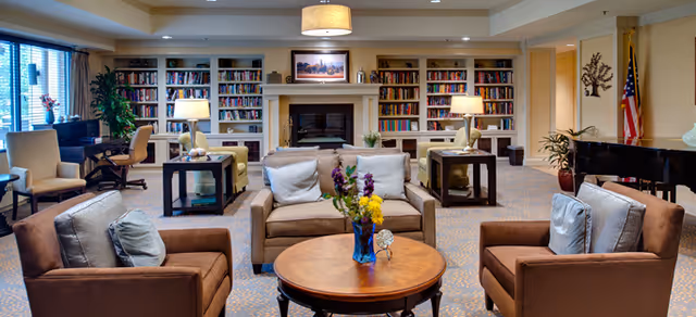A cozy living room area with comfortable armchairs and sofas arranged around a round wooden coffee table with a vase of flowers. The room features built-in bookshelves filled with books, a fireplace with a framed picture above it, table lamps, a piano, and large windows with curtains allowing natural light to enter.