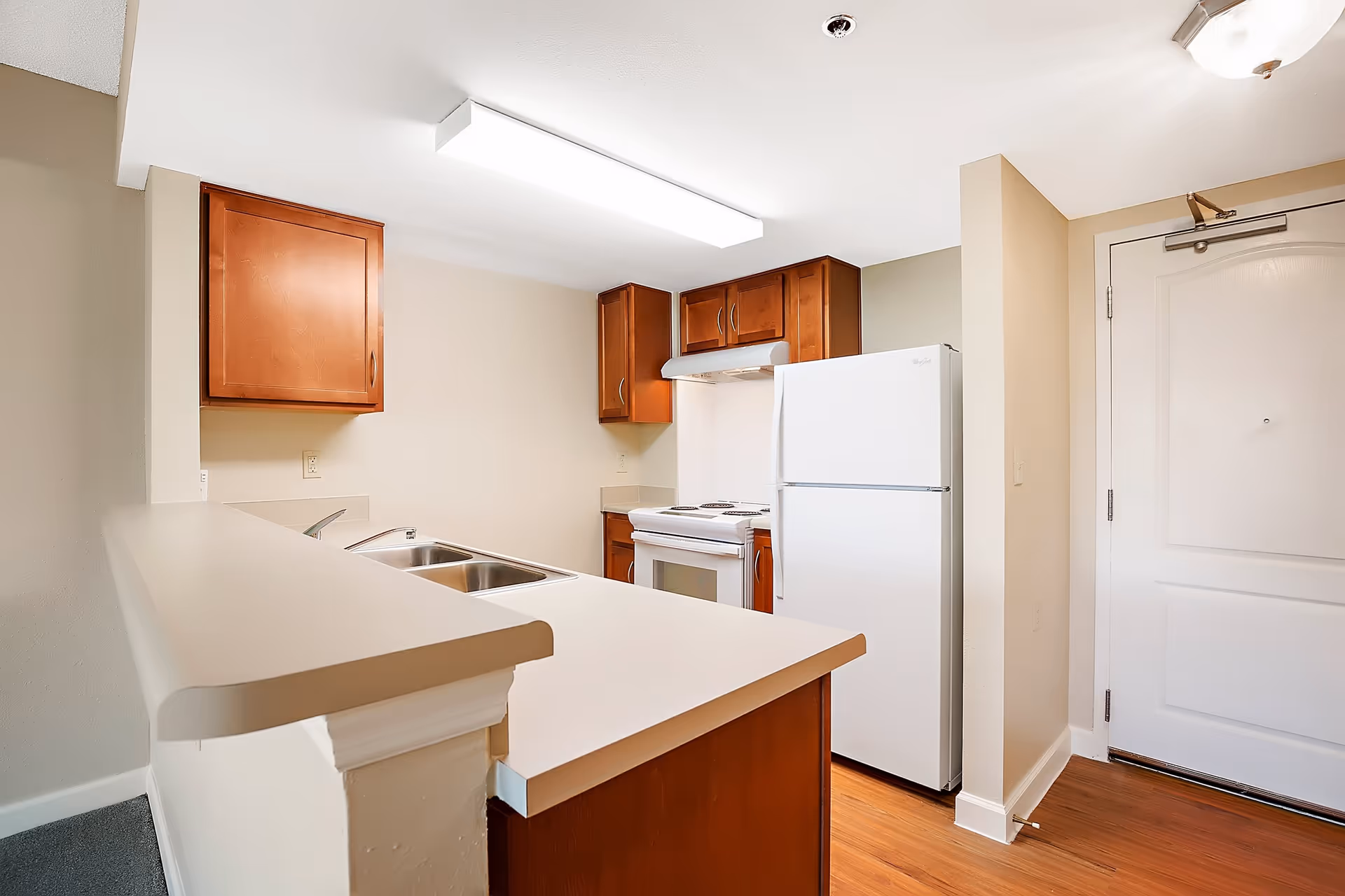 A small kitchen area with wooden cabinets, a white refrigerator, a white stove with an overhead vent, a double sink, and a countertop with a raised bar. The kitchen has beige walls and a wooden floor, with a white door visible to the right.