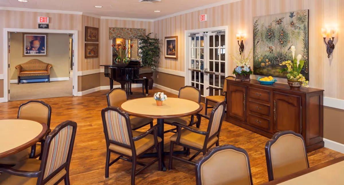 A cozy and well-lit common area in a senior living facility featuring round tables with chairs arranged around them. The room has wooden flooring, beige walls with a chair rail, and decorative wall sconces. A black grand piano is positioned in the corner next to a large plant and a mirror. There is a wooden sideboard with floral arrangements and a large painting above it. Double glass doors and an exit door are visible in the background, along with a small seating area with a cushioned bench.