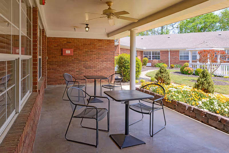 Covered outdoor patio with metal tables and chairs overlooking a landscaped courtyard and brick building.