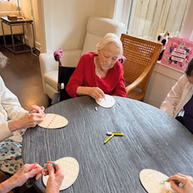 Four elderly individuals sitting around a round table engaged in a craft activity, working on oval-shaped pieces of paper with patterns. The setting appears to be a cozy indoor room with chairs and a window in the background.