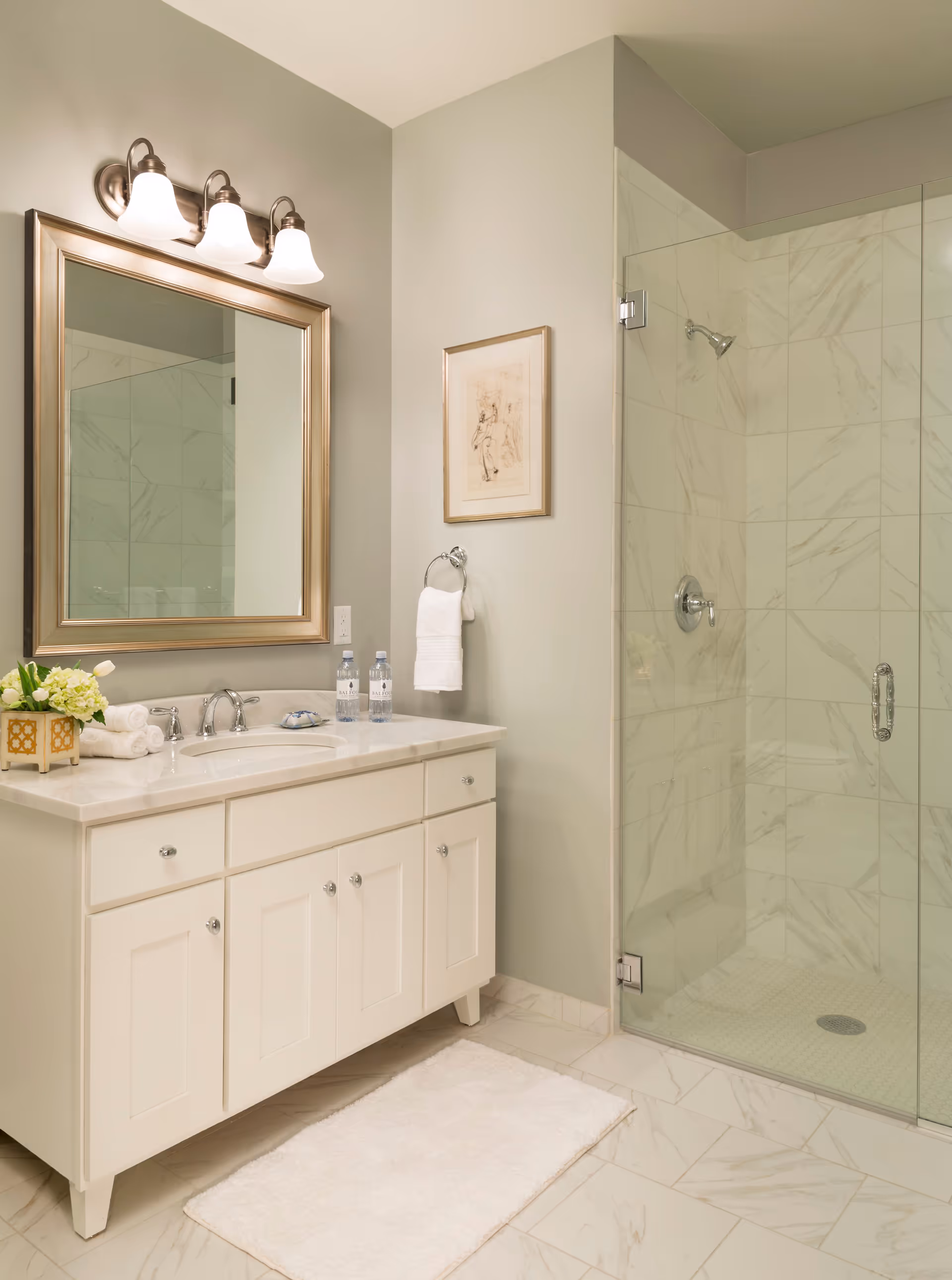 A clean and modern bathroom featuring a white vanity with a marble countertop, a large framed mirror above it, and three light fixtures. There are two bottles of water, a small flower arrangement, and rolled towels on the countertop. To the right is a glass-enclosed shower with marble-patterned tiles. A white bath mat is placed on the floor in front of the vanity, and a towel ring with a white towel hangs on the wall next to a framed artwork.