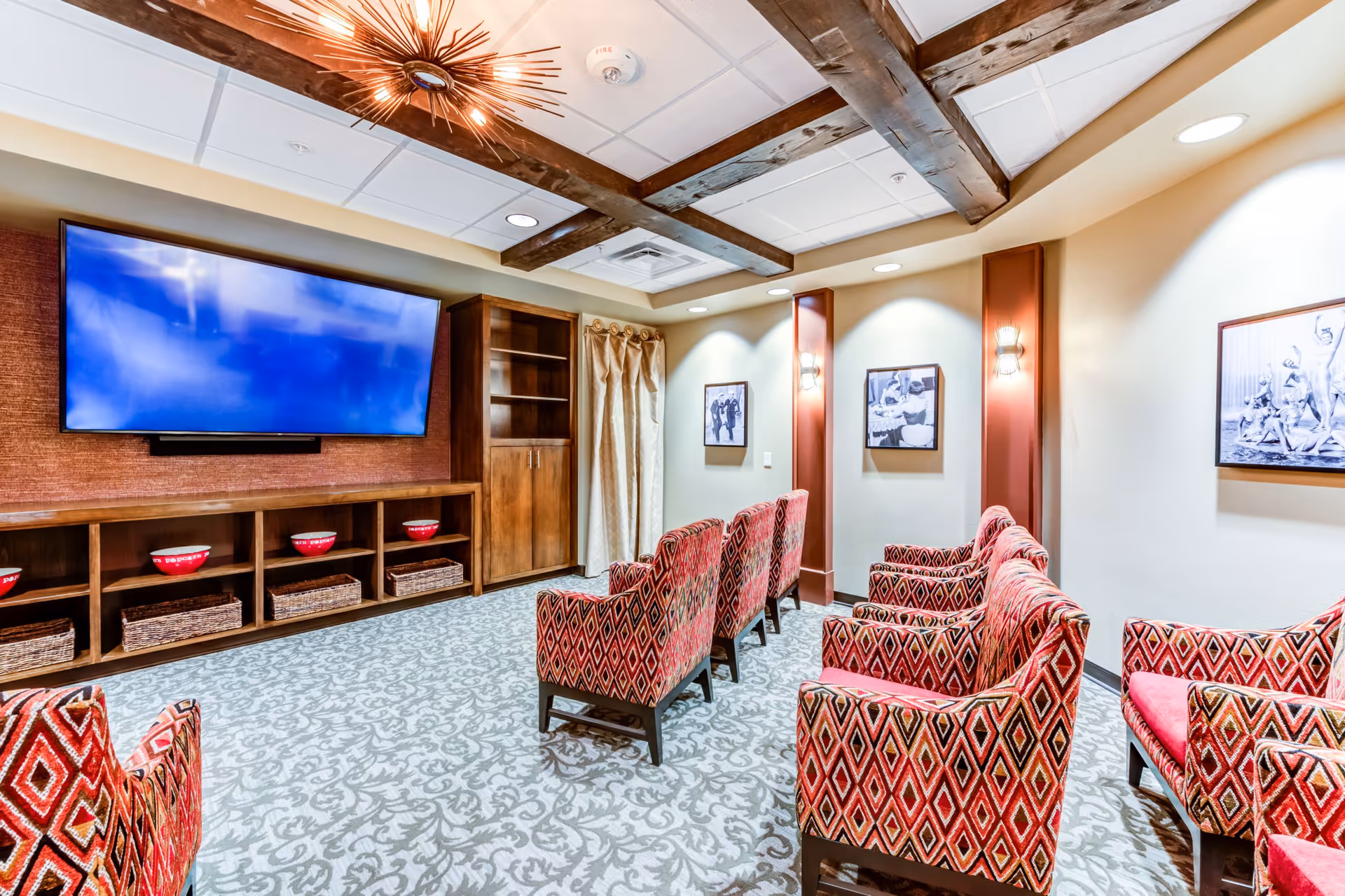 A cozy media room with patterned red armchairs arranged in rows facing a large flat-screen TV mounted on a wooden wall unit. The room features a patterned carpet, wooden ceiling beams, soft lighting, and black-and-white framed photographs on the walls.