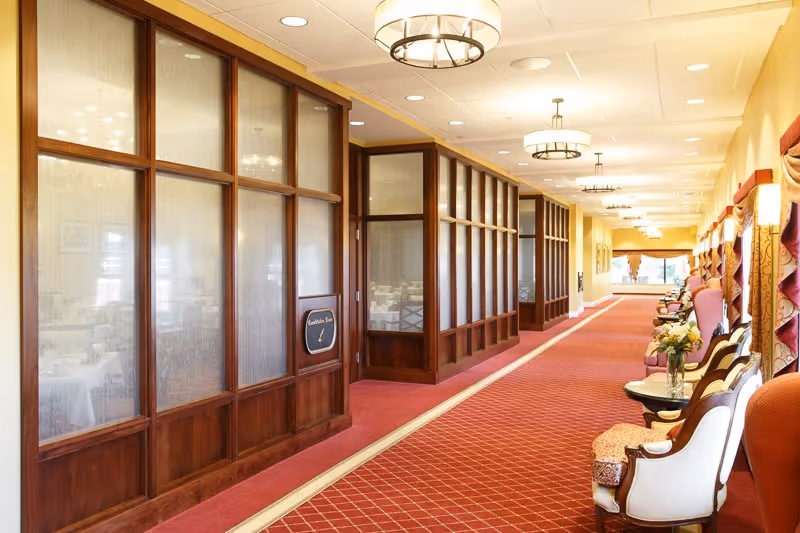 A long, well-lit hallway with a red carpet and wooden framed glass panels on the left side, leading to a dining room. On the right side, there are several upholstered chairs and small tables with flower arrangements, under windows with decorative curtains. Ceiling lights hang evenly spaced along the hallway.