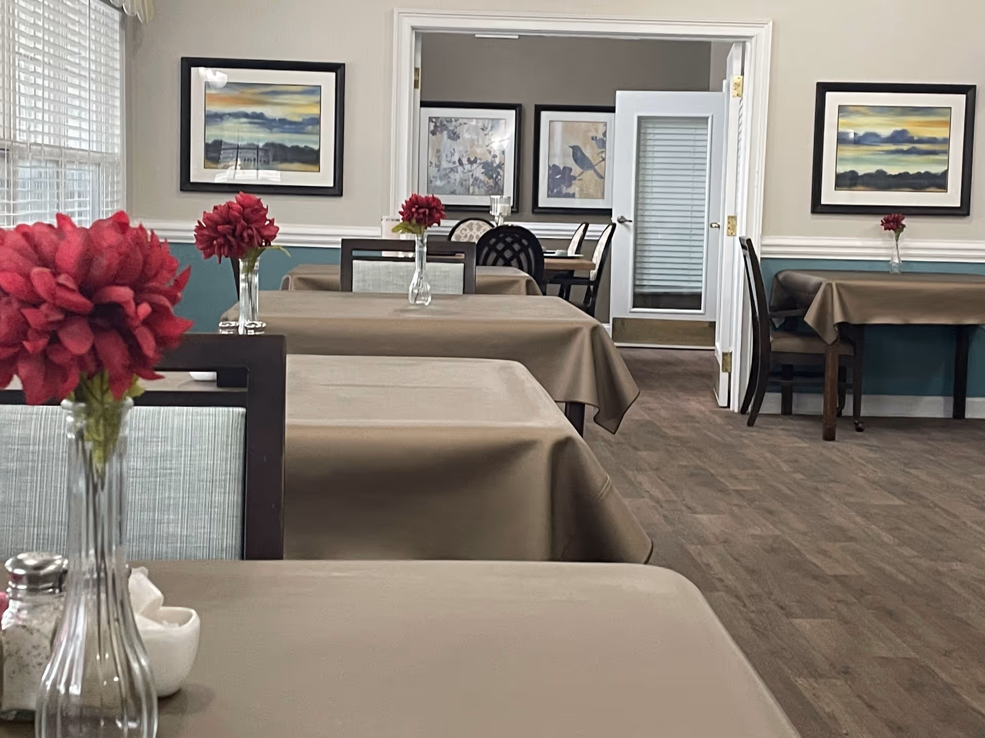 Dining room with rows of tables covered in tan tablecloths, chairs, framed artwork on the walls, and vases holding red flowers.