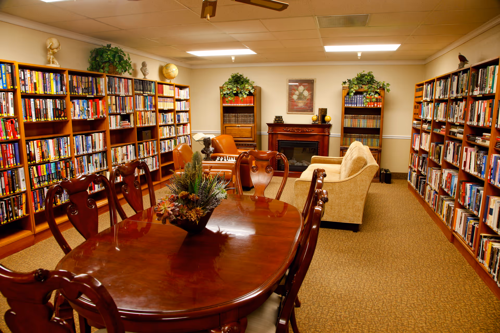 A cozy library-style common room with bookshelves lining the walls, a polished wooden table and chairs, and a seating area with a fireplace.