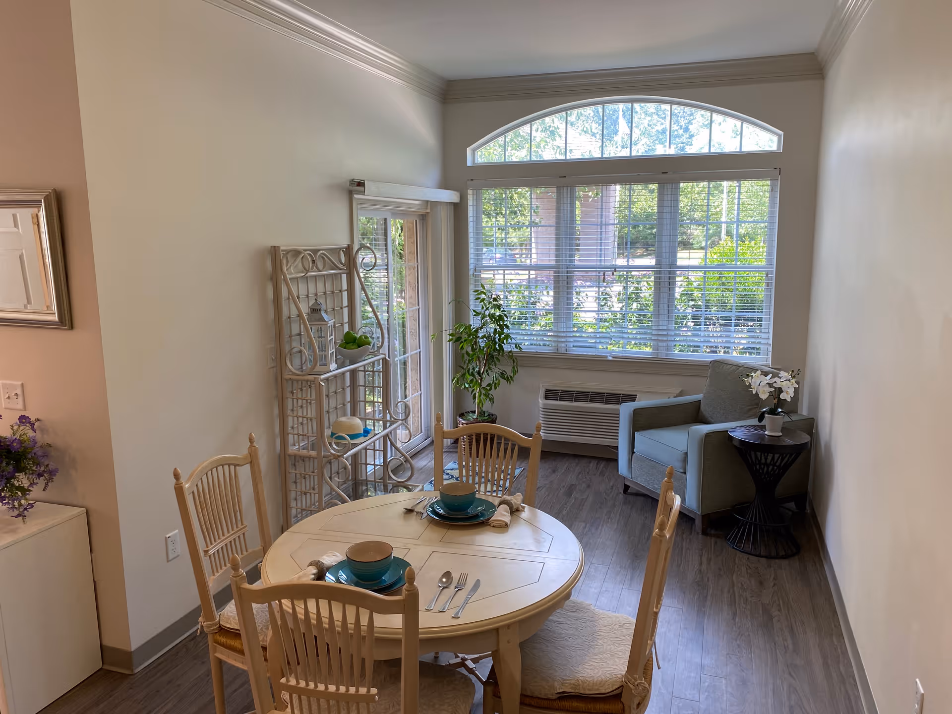 Bright combined dining and sitting area with a round table set for four, an armchair by a large arched window, and decorative shelving.