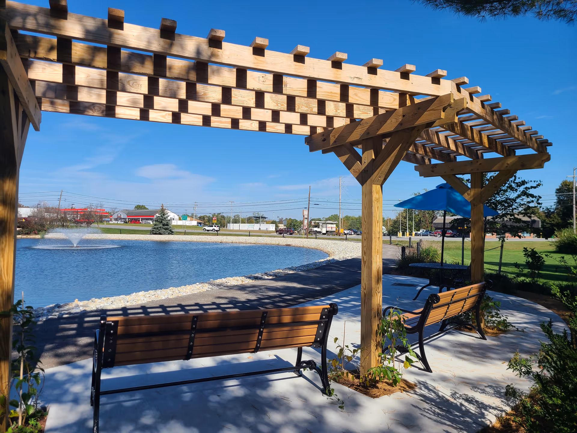 Outdoor seating area with wooden pergola and benches overlooking a pond with a water fountain, a paved walking path, and a blue umbrella-covered table in the background under a clear blue sky.