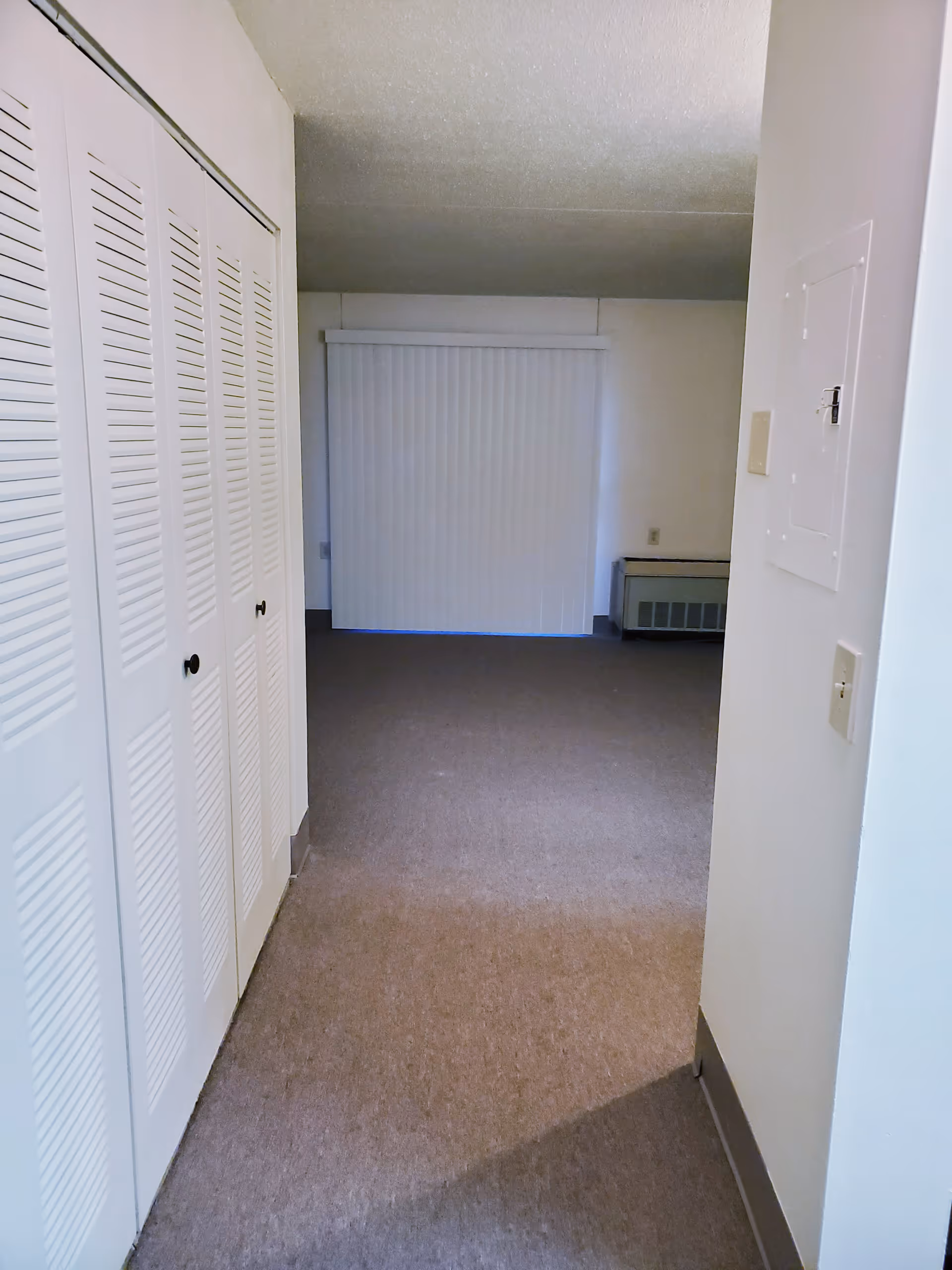 Empty interior room with beige carpet, white walls, a set of white louvered closet doors on the left, a white vertical blind covering a window or sliding door at the far end, and a heating unit below the window.