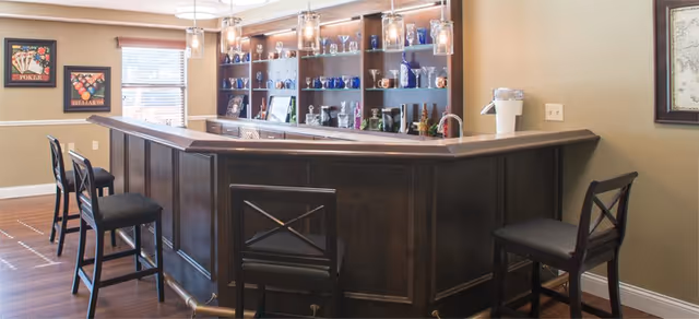 Interior view of a bar area with a dark wooden counter and four high chairs. Behind the bar is a shelving unit displaying various glassware and bottles. The room has beige walls, two windows with blinds, and framed artwork on the walls. Pendant lights hang above the bar counter.