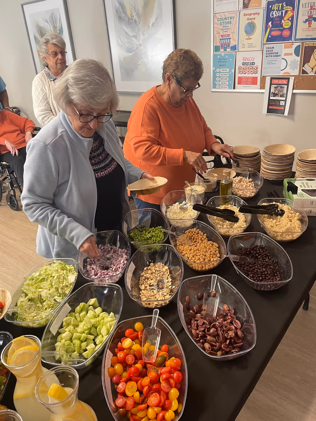 Two elderly women serving themselves food from a salad bar with various fresh ingredients including lettuce, cucumbers, cherry tomatoes, beans, chickpeas, and onions. One woman is wearing a light blue jacket and glasses, while the other is in an orange sweater. In the background, there are other elderly people and a bulletin board with colorful flyers and announcements.