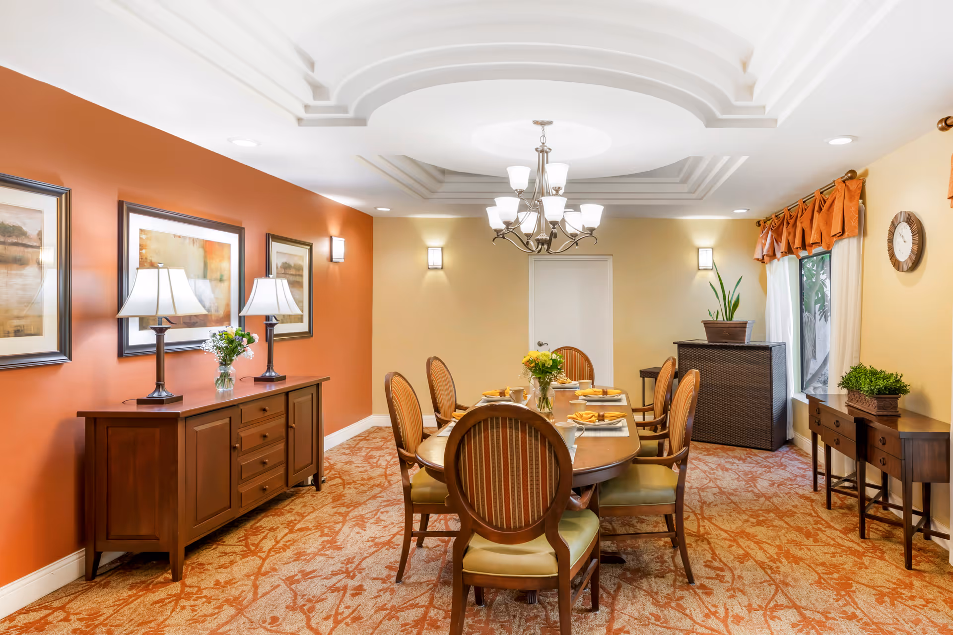 Well-lit dining room with a round table set for six, wooden sideboards, framed artwork, and a central chandelier.