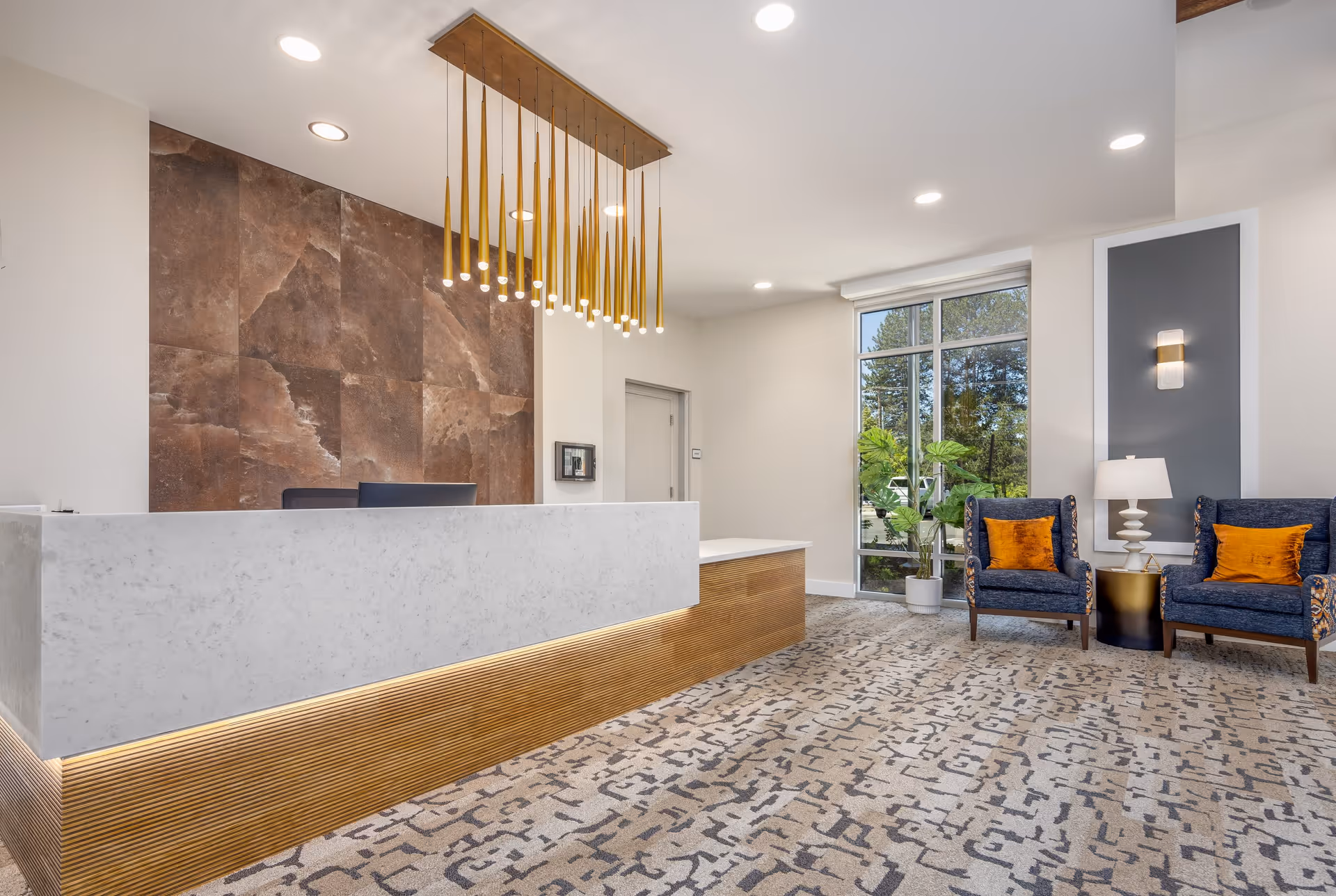 A modern reception area with a white marble front desk featuring wooden paneling at the base. Behind the desk is a wall with large brown textured tiles. Above the desk hangs a contemporary light fixture with multiple gold pendant lights. To the right, there are two blue armchairs with orange cushions, a small round black table with a white lamp, and a large window showing greenery outside. The floor is covered with a patterned carpet in neutral tones.