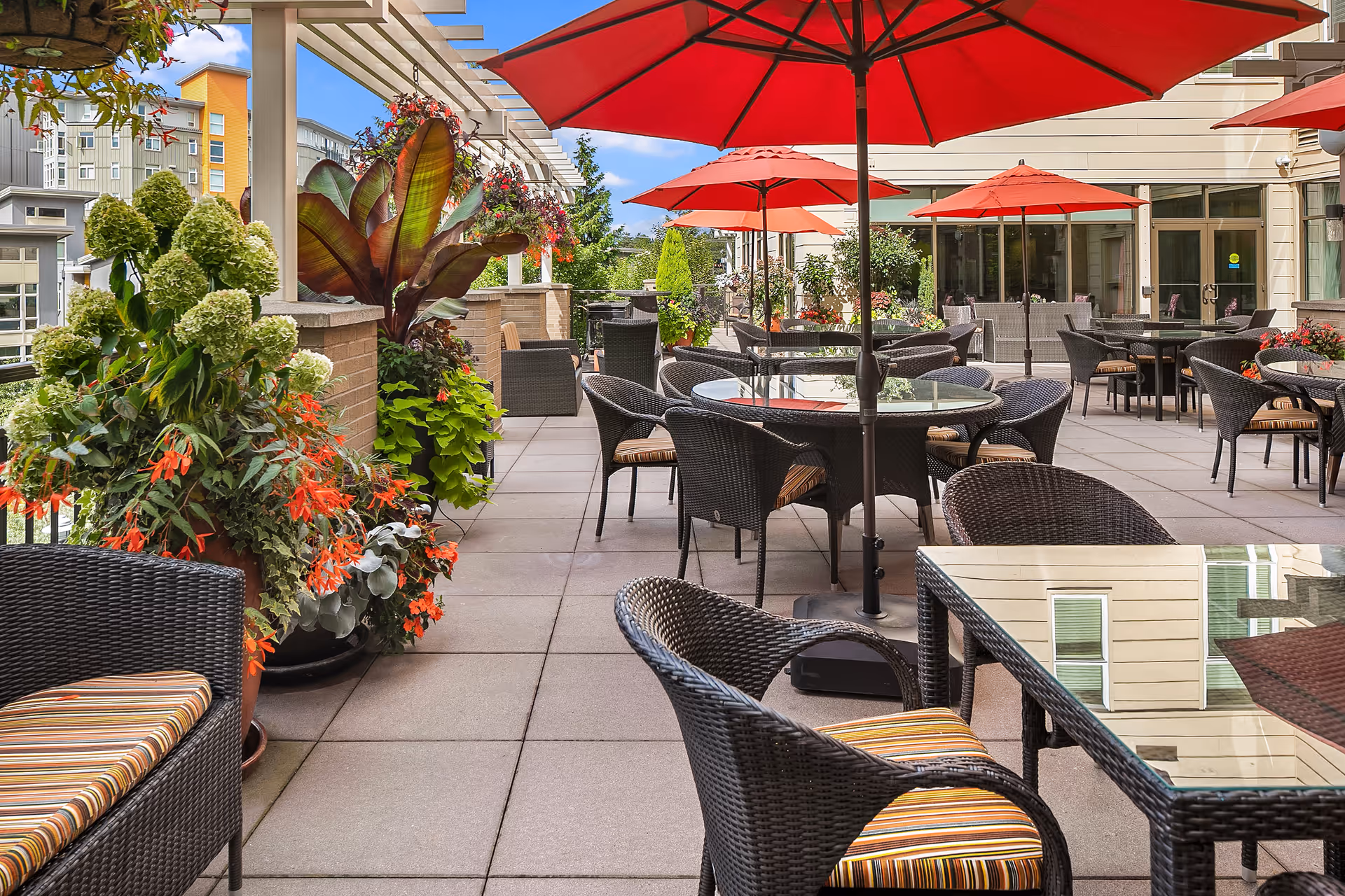 Outdoor patio area at Aljoya Thornton Place with multiple round glass-top tables surrounded by wicker chairs with striped cushions. Several red umbrellas provide shade. The patio is decorated with large potted plants and flowers, and buildings are visible in the background under a blue sky with some clouds.