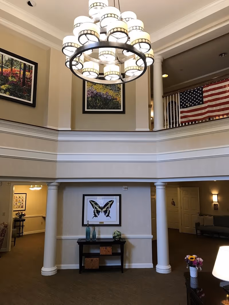 Interior view of a senior living facility lobby with a large circular chandelier hanging from the ceiling. The space features two white columns, framed artwork including a butterfly print, and an American flag hanging on the upper level railing. There is a small table with decorative items and a vase of flowers on a nearby surface.