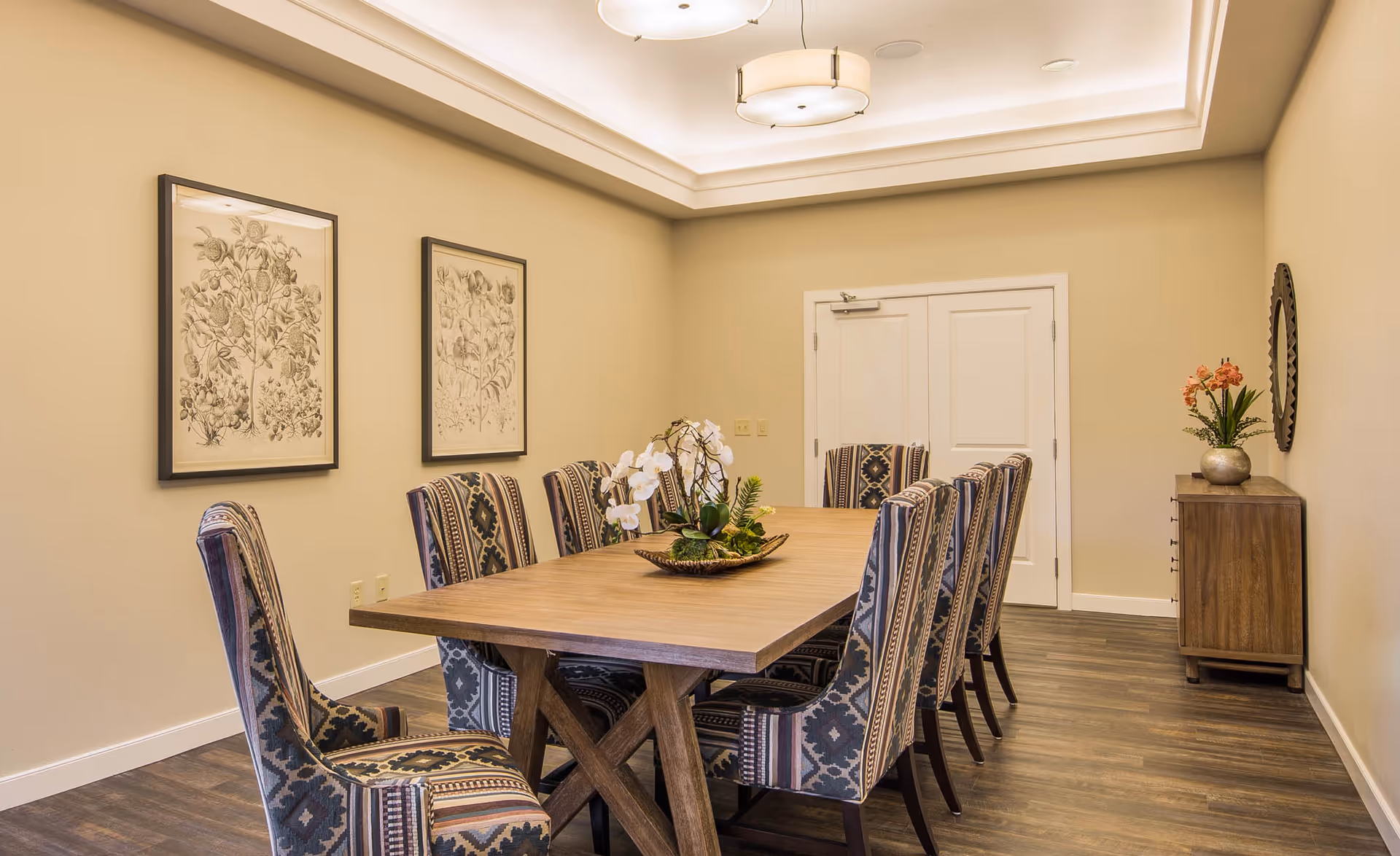 A dining room with a long wooden table surrounded by patterned upholstered chairs, neutral walls, framed botanical prints, and floral centerpieces.