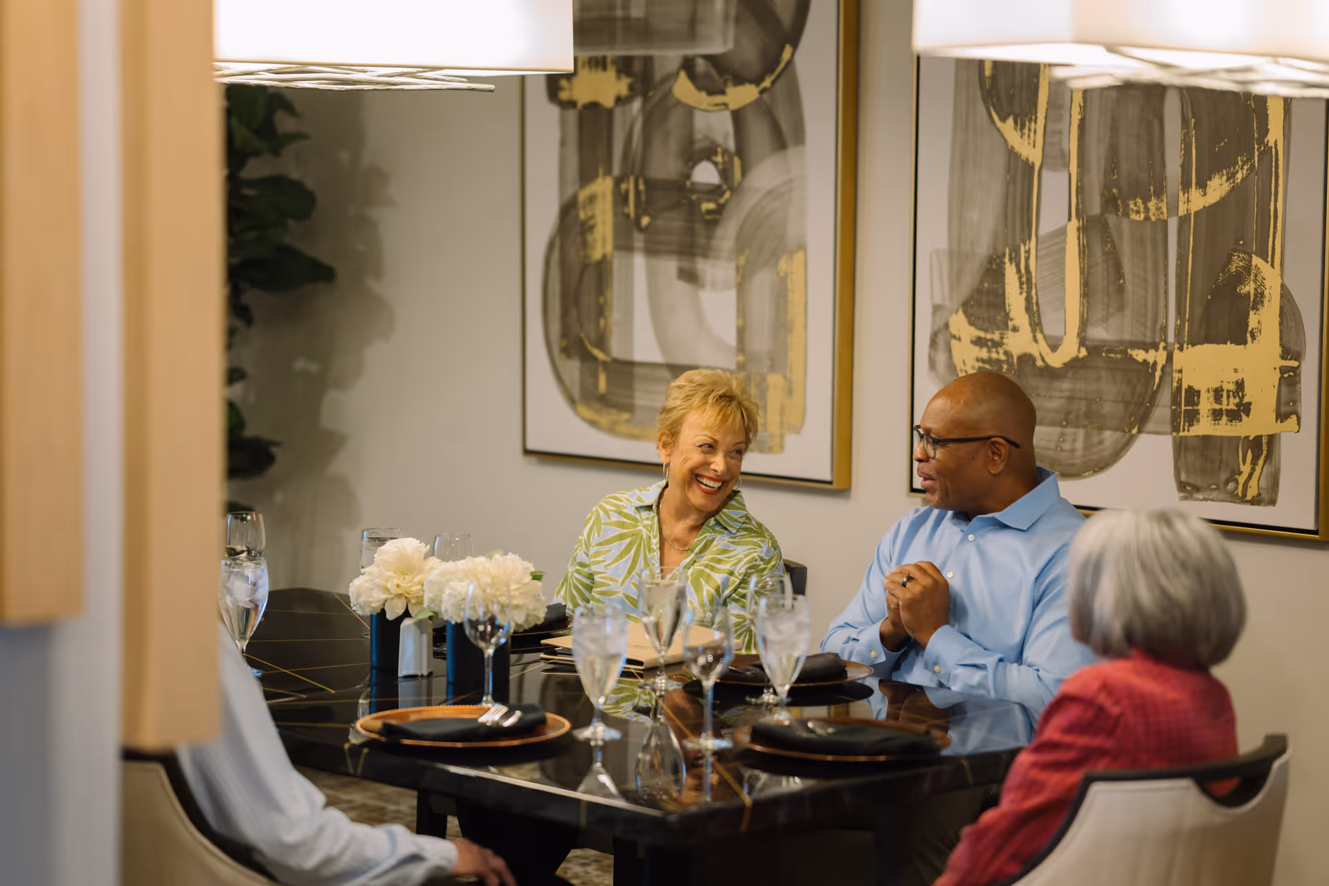Three elderly people sitting around a dining table engaged in conversation. The table is set with plates, glasses, and a centerpiece of white flowers. Abstract artwork is visible on the wall behind them.