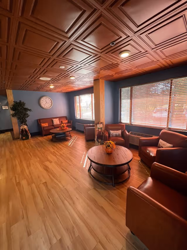 A cozy living room area with wooden flooring and a decorative wooden ceiling. The room features several brown leather armchairs and a matching sofa arranged around two round wooden coffee tables with flower arrangements. Large windows with blinds allow natural light to fill the space. A wall clock and a potted plant are visible in the background.