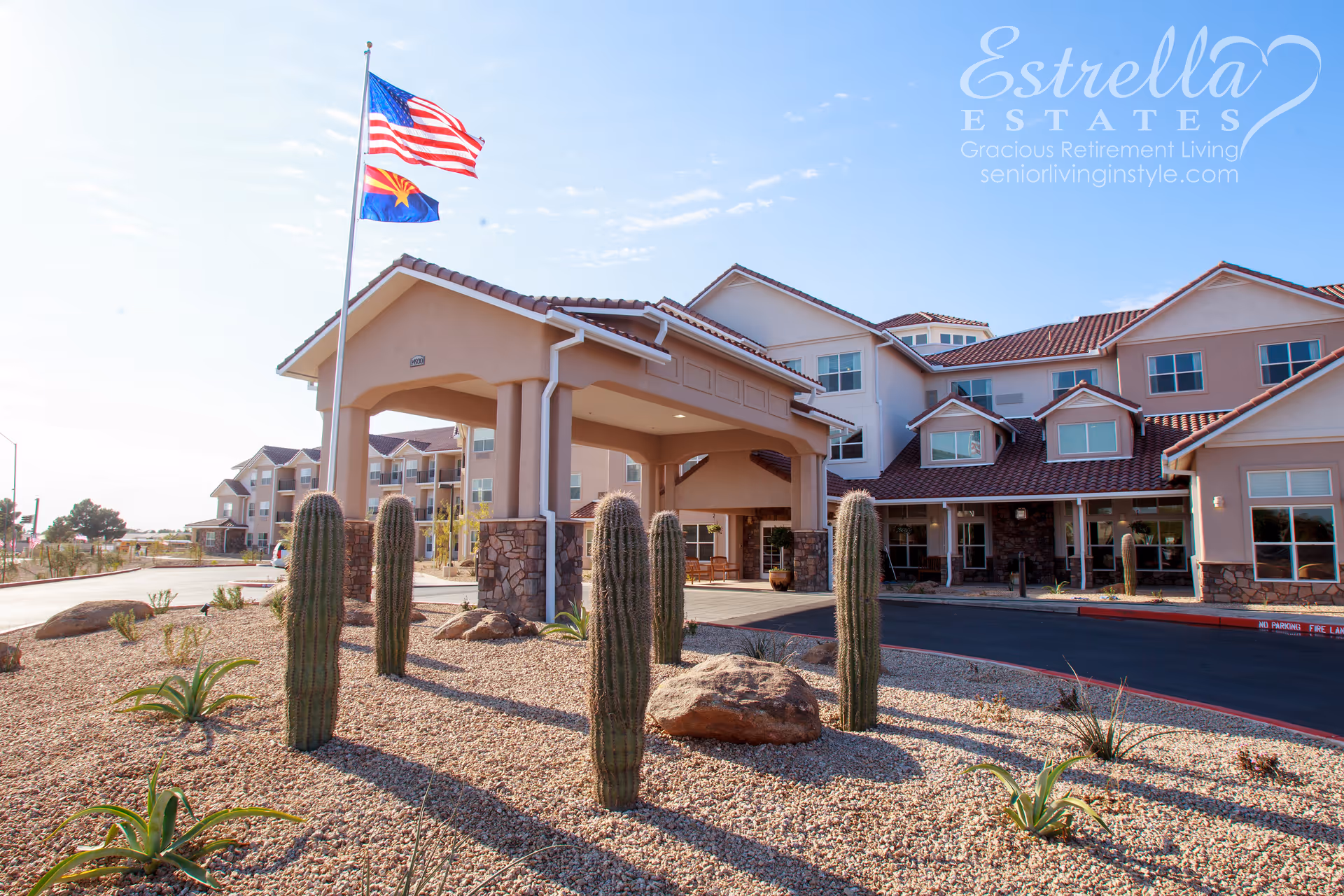 Exterior view of Estrella Estates Gracious Retirement Living facility with a covered entrance, desert landscaping featuring tall cacti and rocks, and two flags flying on a flagpole against a clear blue sky.