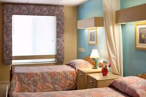 Pastel-walled hotel-style bedroom with two beds, nightstand, lamp, and curtains.