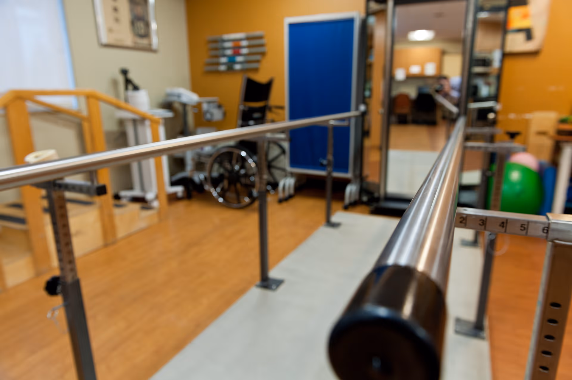 Physical therapy room with parallel bars for walking exercises, a wheelchair, exercise balls, and other rehabilitation equipment in a healthcare facility.