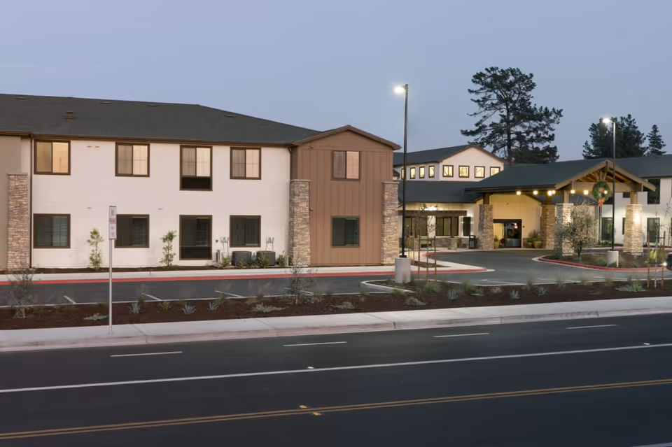 Front exterior of a two-story senior living facility with an illuminated covered entrance, driveway, and landscaped parking area at dusk.