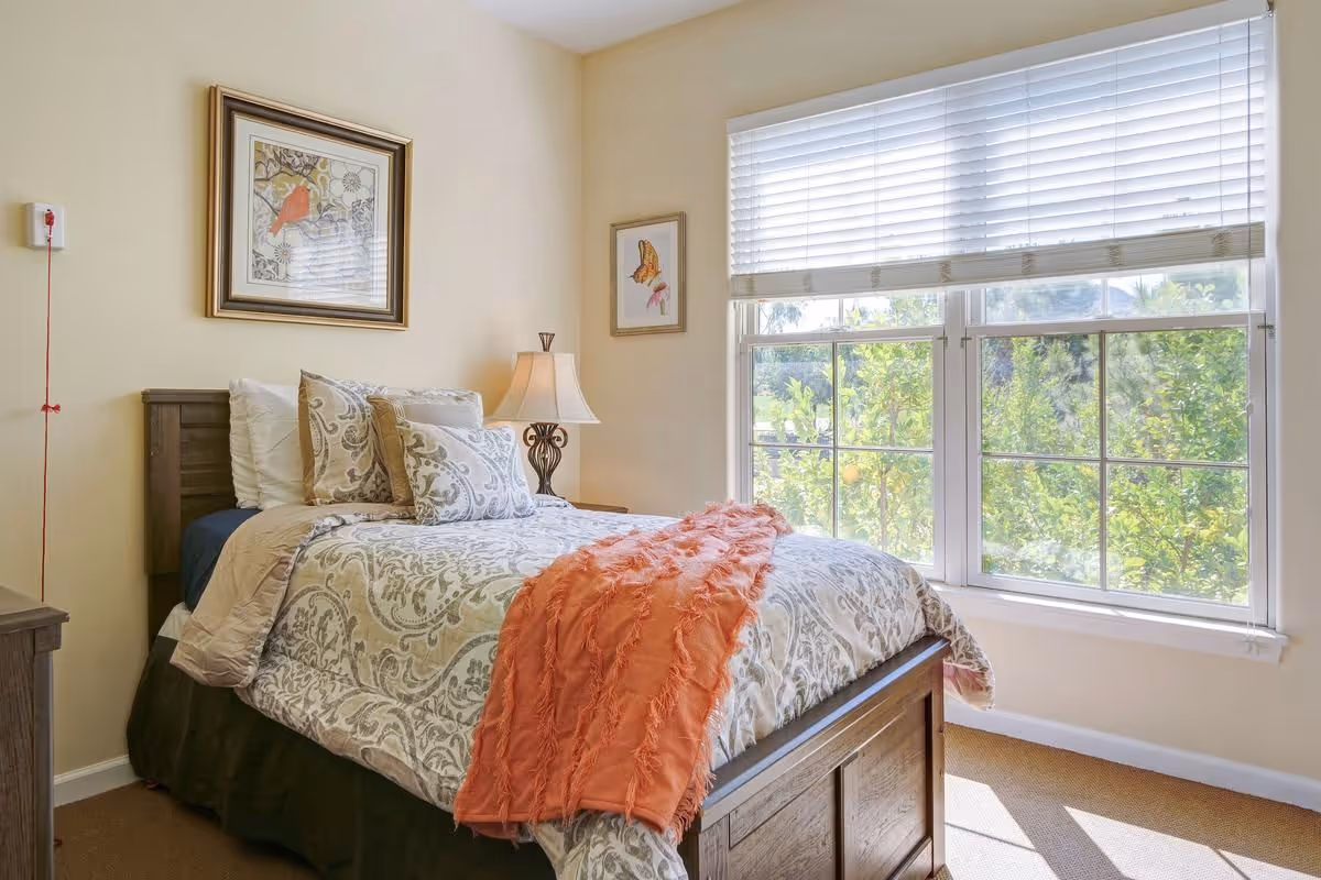 A cozy bedroom with a wooden bed frame, patterned bedding, and an orange throw blanket. There are two framed pictures on the walls, a bedside table with a lamp, and a large window with white blinds letting in natural light and showing greenery outside.