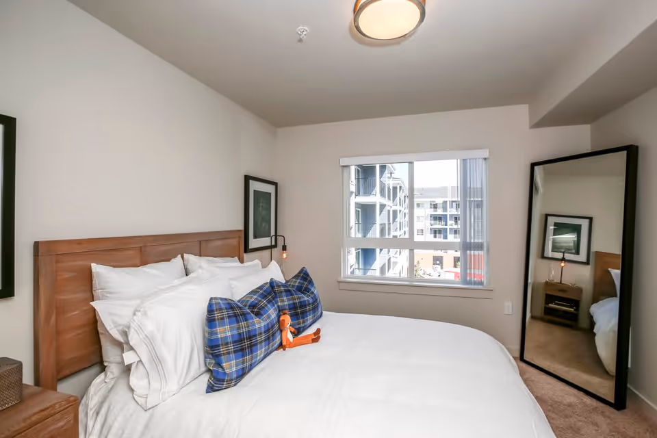 A bedroom with a wooden headboard bed made with white linens and blue plaid pillows. A small orange stuffed toy is placed on the bed. There is a large window with vertical blinds showing an exterior view of nearby buildings. A full-length mirror is leaning against the wall to the right, reflecting part of the bed and a framed picture on the wall. The walls are painted light beige and the floor is carpeted.