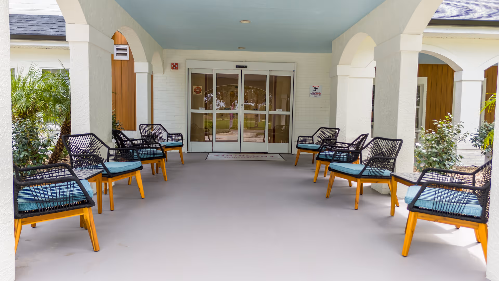 Covered outdoor seating area with black wicker chairs featuring blue cushions arranged along both sides of a walkway leading to glass double doors. The area is framed by white columns and arches, with greenery visible on the sides.