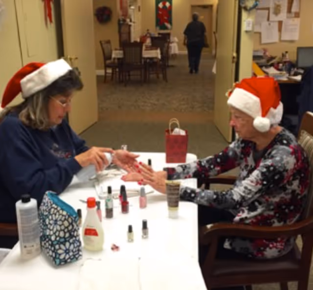 Two women wearing Santa hats sit at a table in a senior facility while one gives the other a manicure.