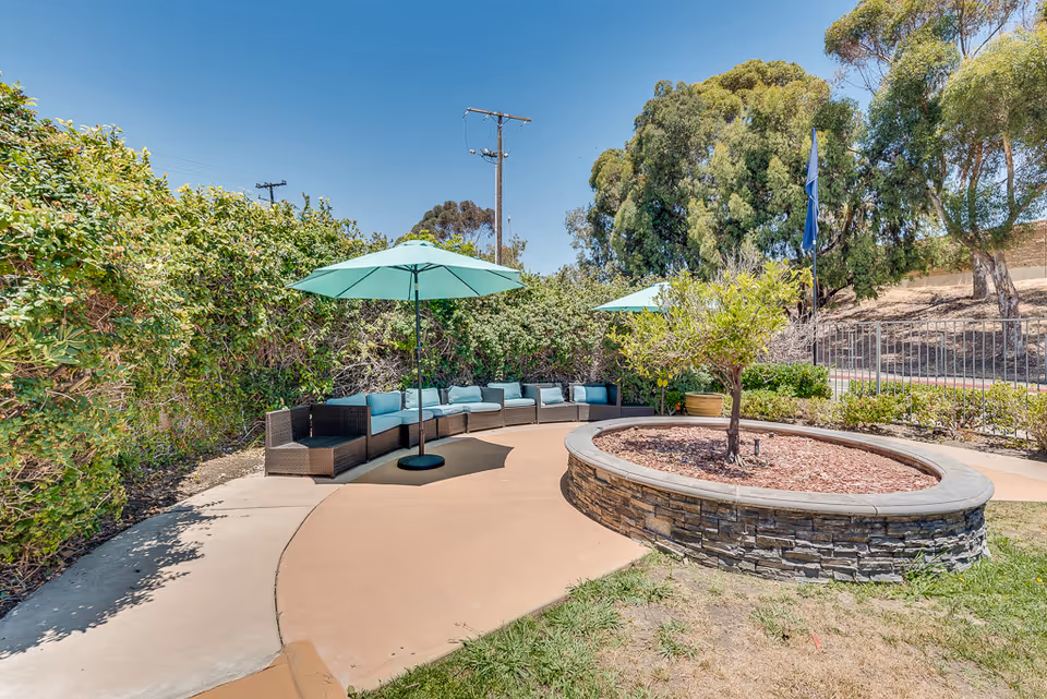 Outdoor seating area with curved wicker sofa and blue cushions under two large blue umbrellas, surrounded by greenery and trees on a sunny day.