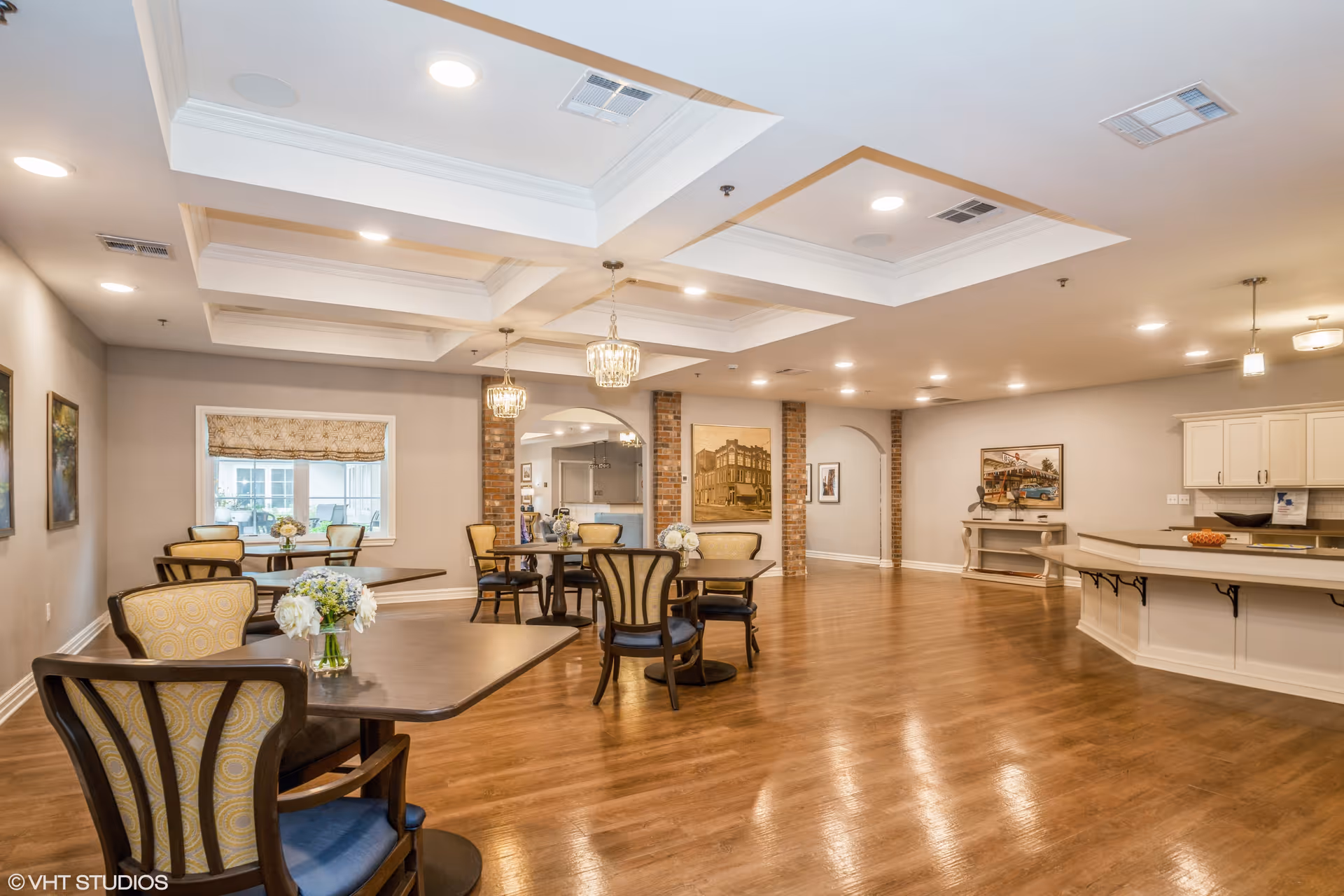 A spacious dining room with wooden floors and several tables and chairs arranged neatly. The ceiling features recessed lighting and decorative tray ceilings with hanging chandeliers. The walls are adorned with framed artwork and there is a window with a patterned Roman shade. A kitchen counter with white cabinetry is visible on the right side of the room.