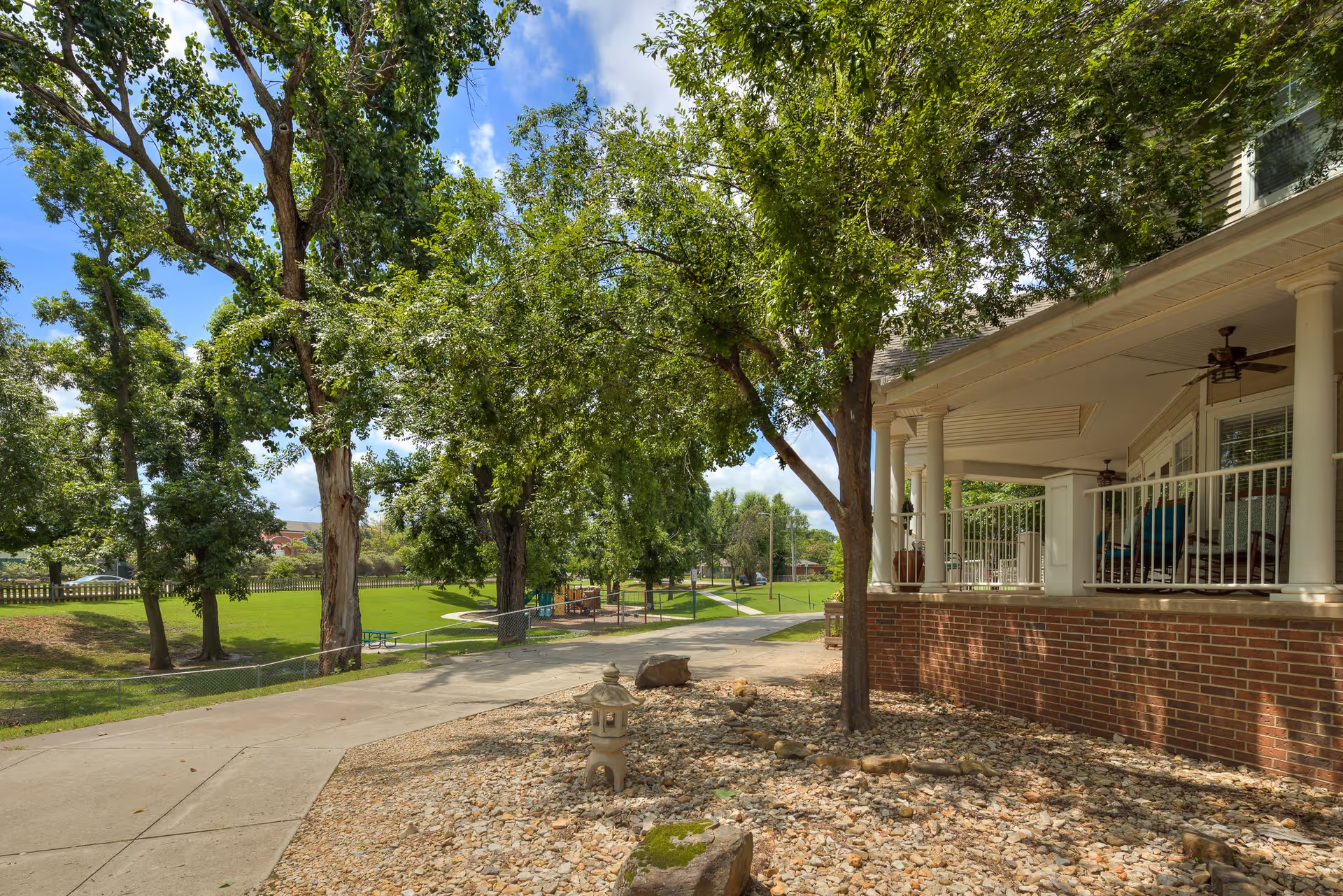 Covered porch and brick exterior overlooking a tree-lined walkway, grassy lawn, and distant playground.