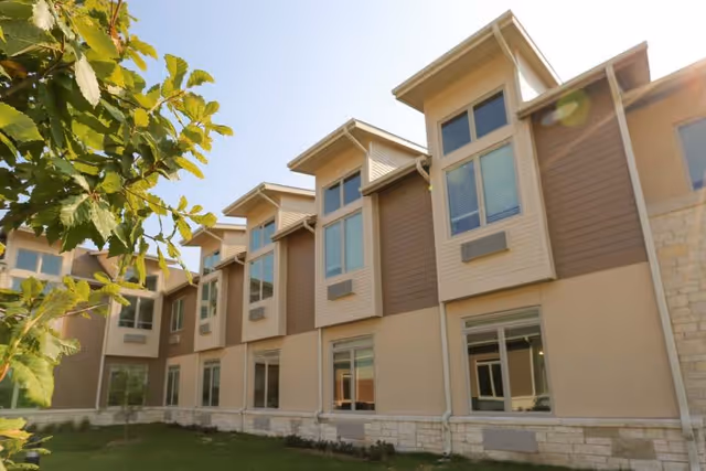 Exterior view of a two-story building with multiple windows, beige and brown siding, and a stone foundation. There is a small grassy area with a tree in the foreground under a clear blue sky.