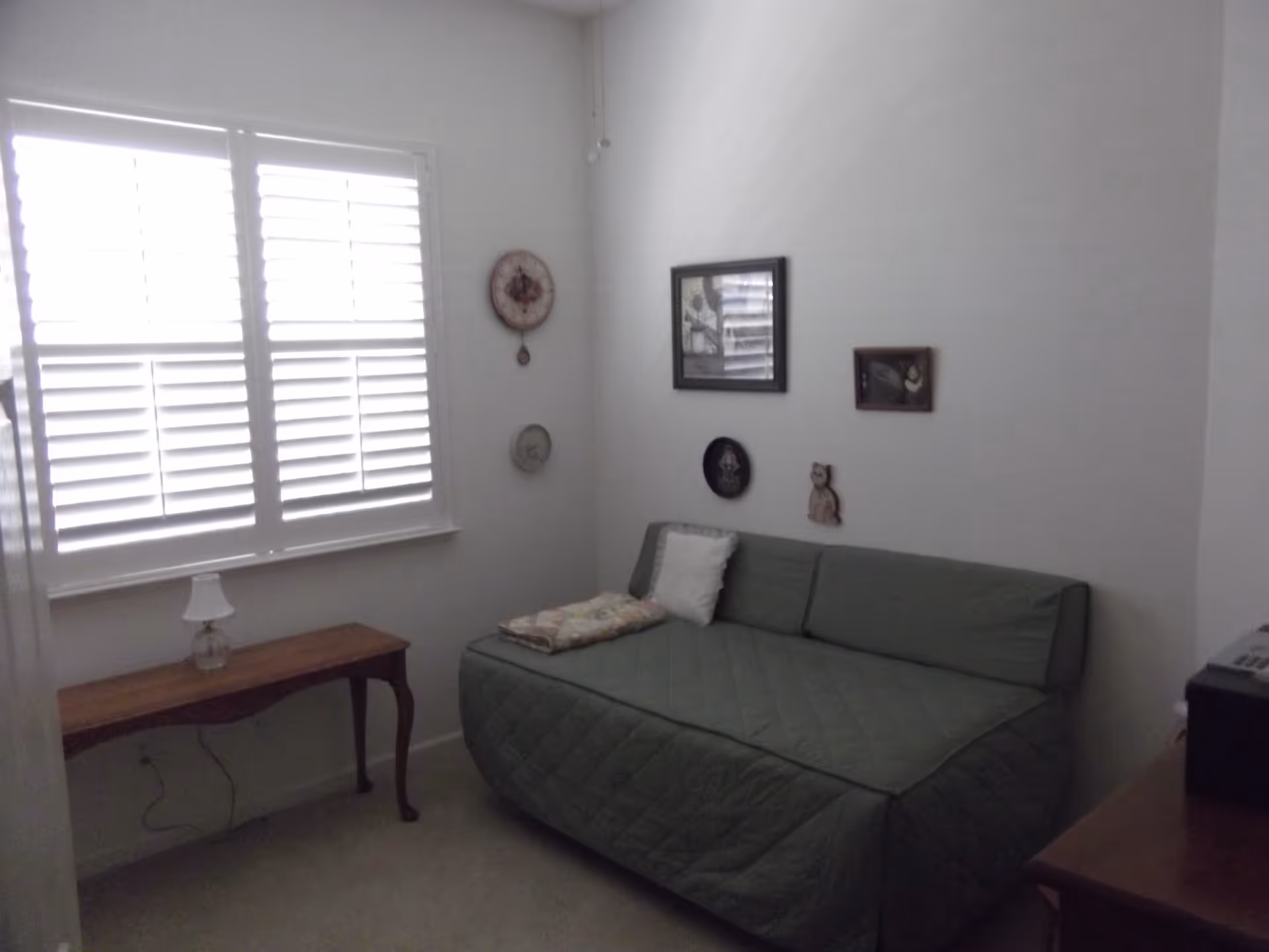A small bedroom with a green daybed covered with a quilt and a white pillow. There is a wooden side table with a small lamp next to a window with white plantation shutters. The walls are white and decorated with a few small framed pictures and decorative plates.