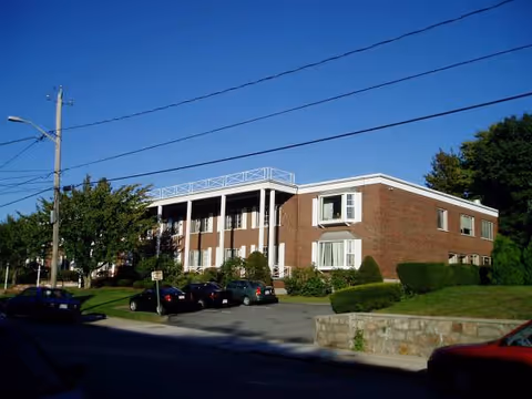 Brick two-story senior living center exterior with parked cars and blue sky.