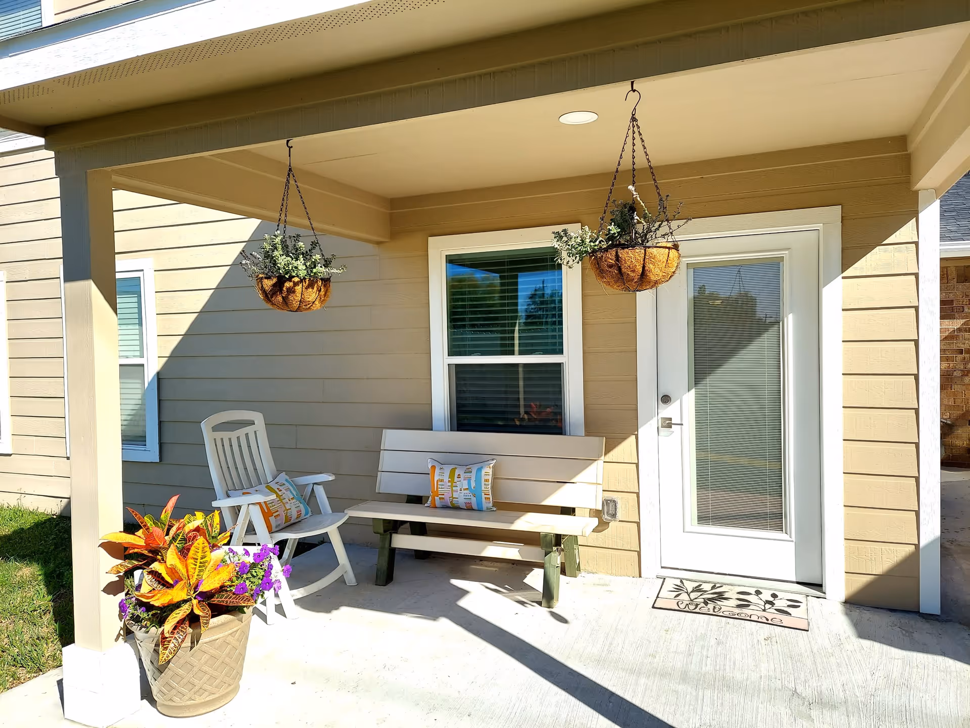 Covered entry porch with a bench, white chair, potted and hanging plants, and a glass door to the house.