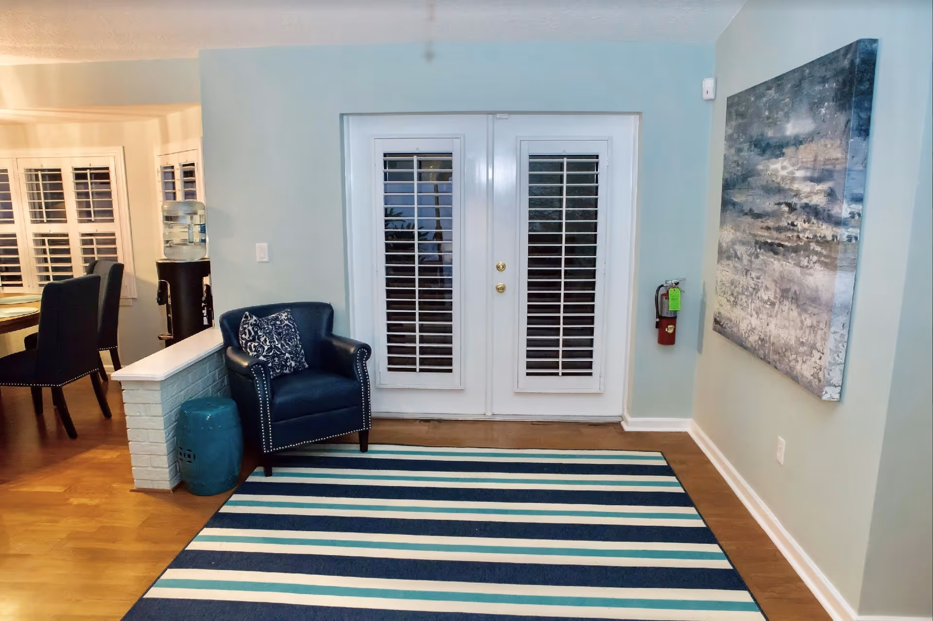 Interior view of a room with light blue walls featuring a set of white double doors with glass panes and white shutters. In front of the doors is a striped rug in shades of blue and white. To the left, there is a dark blue armchair with a patterned cushion and a small blue ceramic stool next to it. A white half-wall separates this area from a dining space with a wooden table and black chairs. A water dispenser is visible near the dining area. On the right wall, there is a large abstract painting in shades of blue, gray, and white. A fire extinguisher is mounted on the wall below the painting.