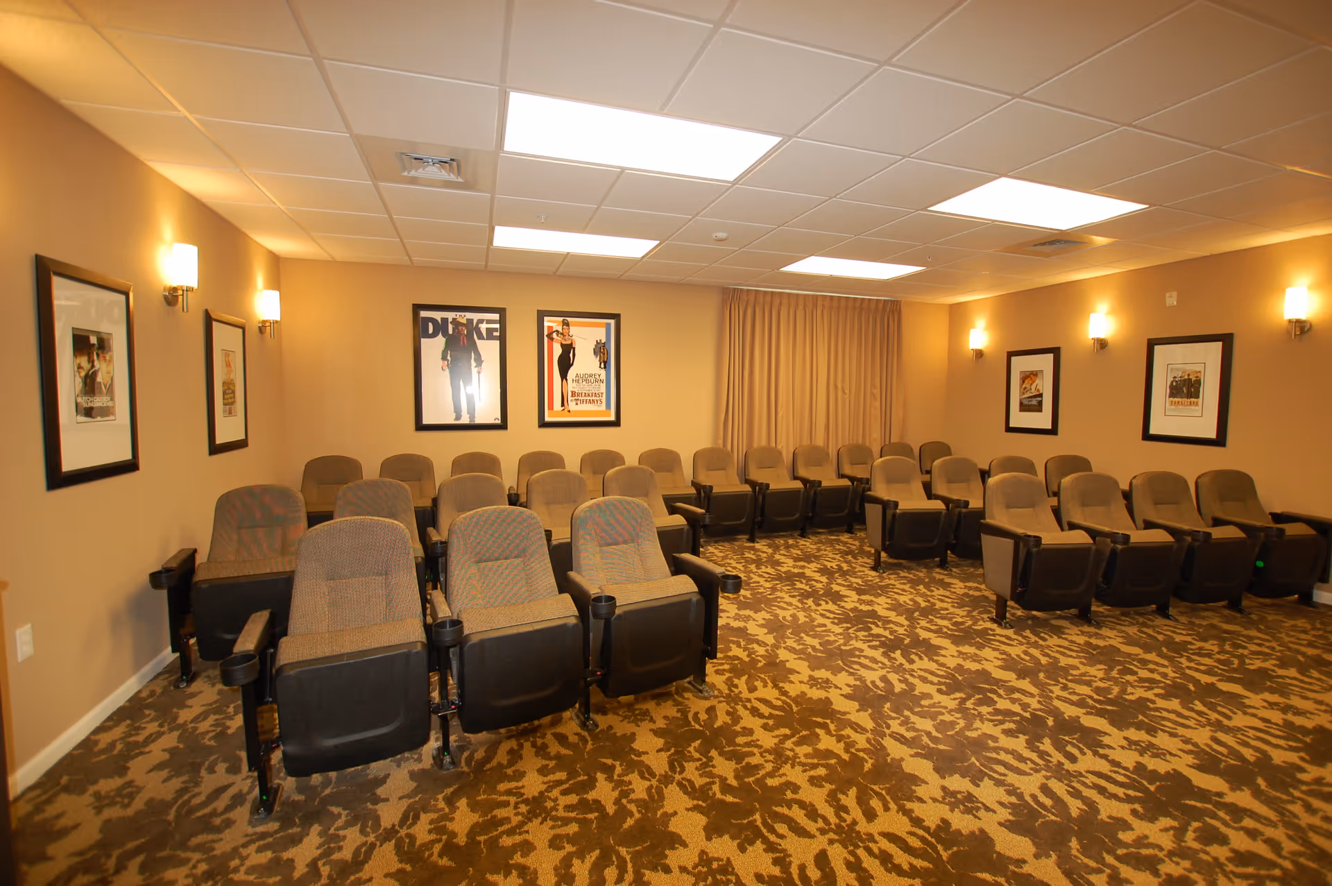 A small theater room with three rows of cushioned seats arranged in a semi-circle facing the front. The walls are beige with framed movie posters and wall sconces providing warm lighting. The floor is carpeted with a patterned design, and a curtain covers the front wall.