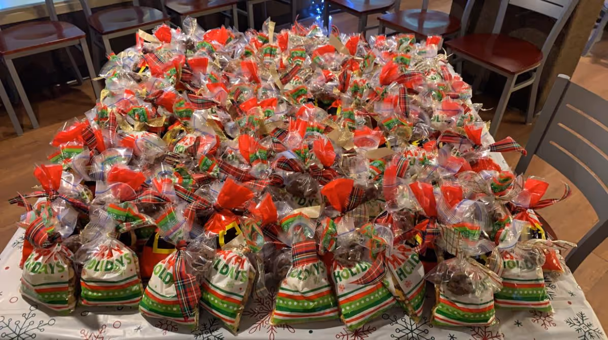 A large table covered with numerous holiday-themed gift bags filled with treats, each bag decorated with festive colors like red, green, and white, and tied with ribbons. The table is set in a room with wooden chairs and a wooden floor.