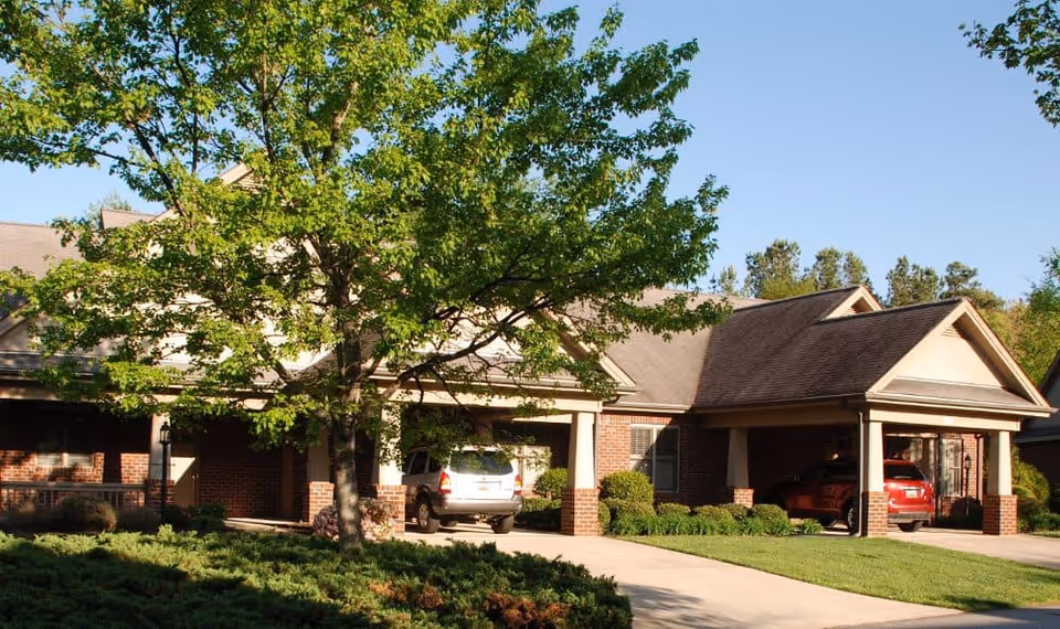Exterior view of a single-story brick building with a covered driveway and two parked cars, surrounded by green trees and shrubs under a clear blue sky.