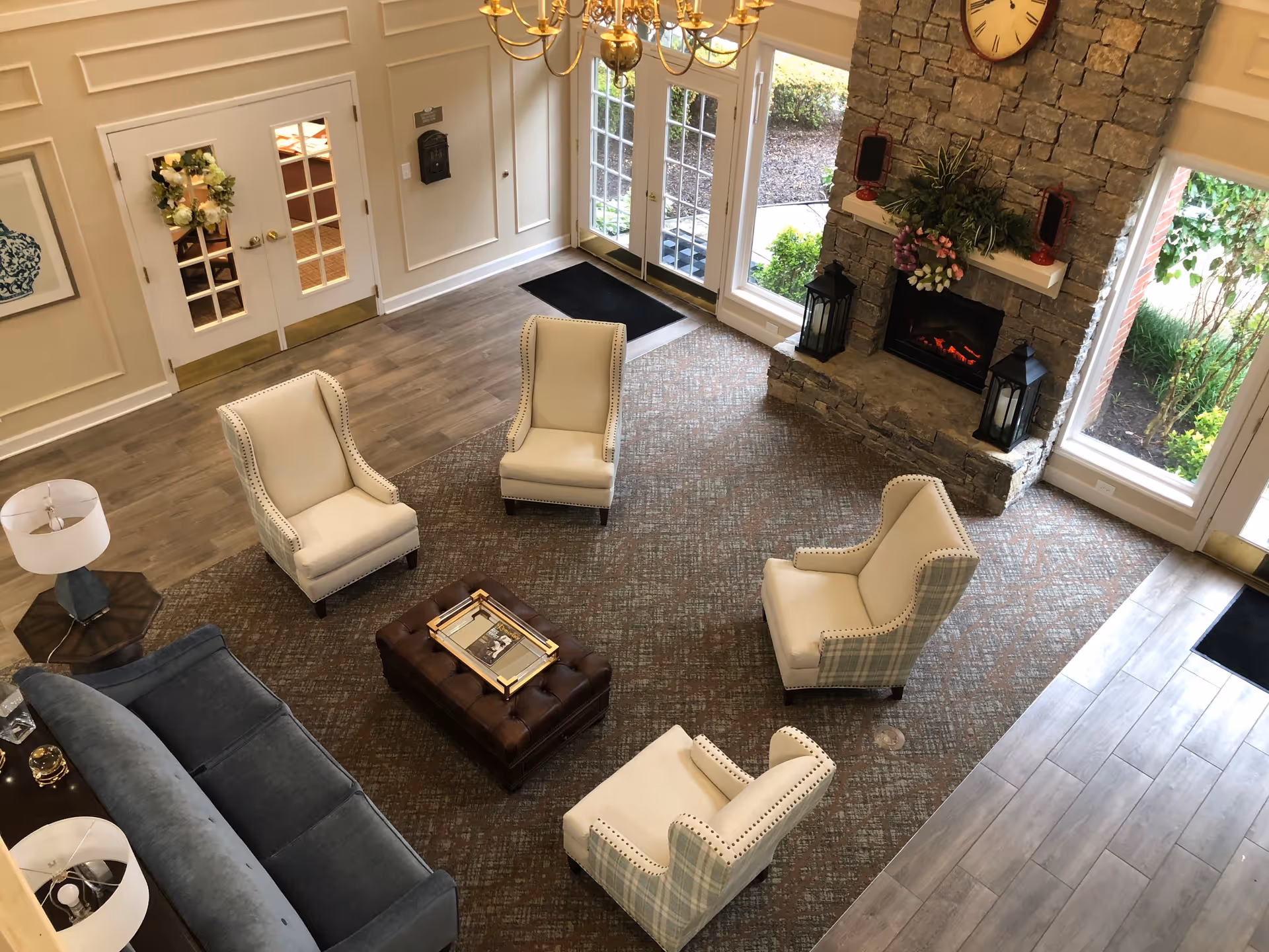 A cozy living room area with four beige armchairs arranged around a brown tufted ottoman. There is a blue sofa on the left side and a wooden side table with a lamp. The room features a stone fireplace with a clock above it and decorative plants and lanterns on the mantel. Large windows and glass doors let in natural light and show greenery outside. The floor is a combination of wood and carpet.