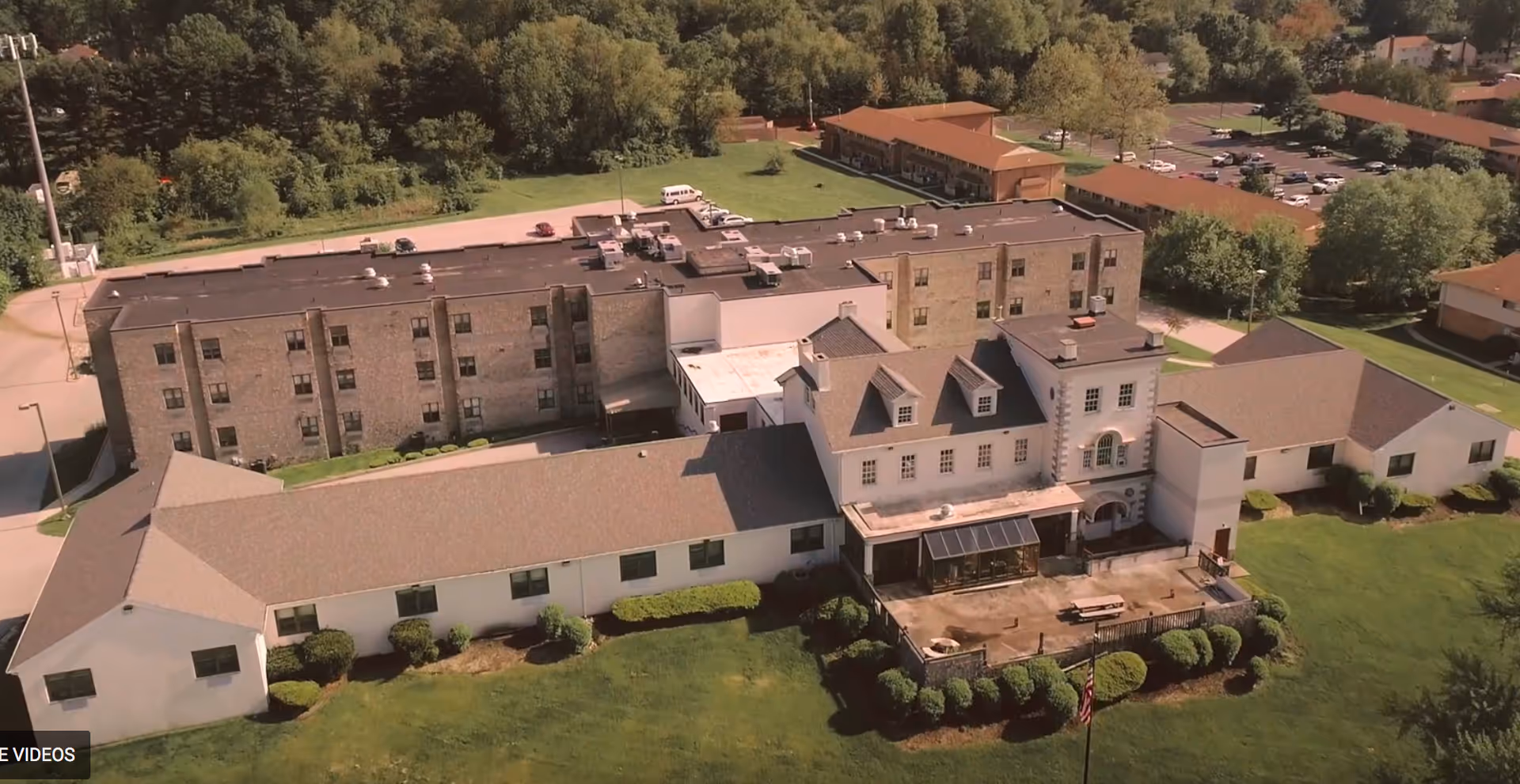 Aerial view of a senior living facility named Aventura At Pembrooke, showing multiple connected buildings with a mix of brick and white exterior walls, surrounded by green lawns, trees, and parking areas.