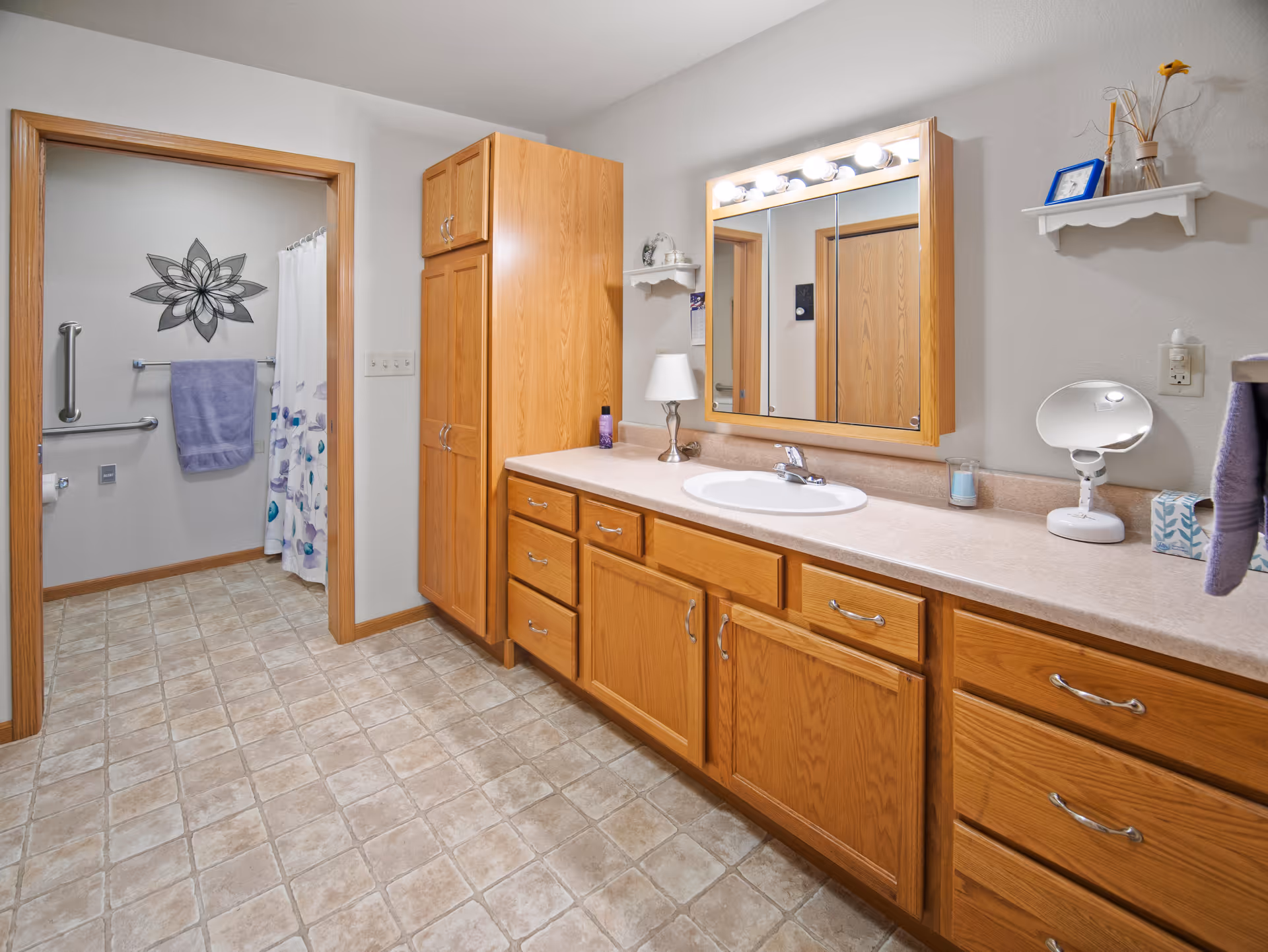 A spacious bathroom with a large wooden vanity featuring multiple drawers and cabinets, a countertop with a sink, a mirror with lights above it, and a small lamp. The bathroom has tiled flooring and light-colored walls. In the background, there is a doorway leading to a shower area with a grab bar, a towel rack holding a purple towel, and a shower curtain with a floral pattern. A decorative flower wall art is visible on the wall inside the shower area.