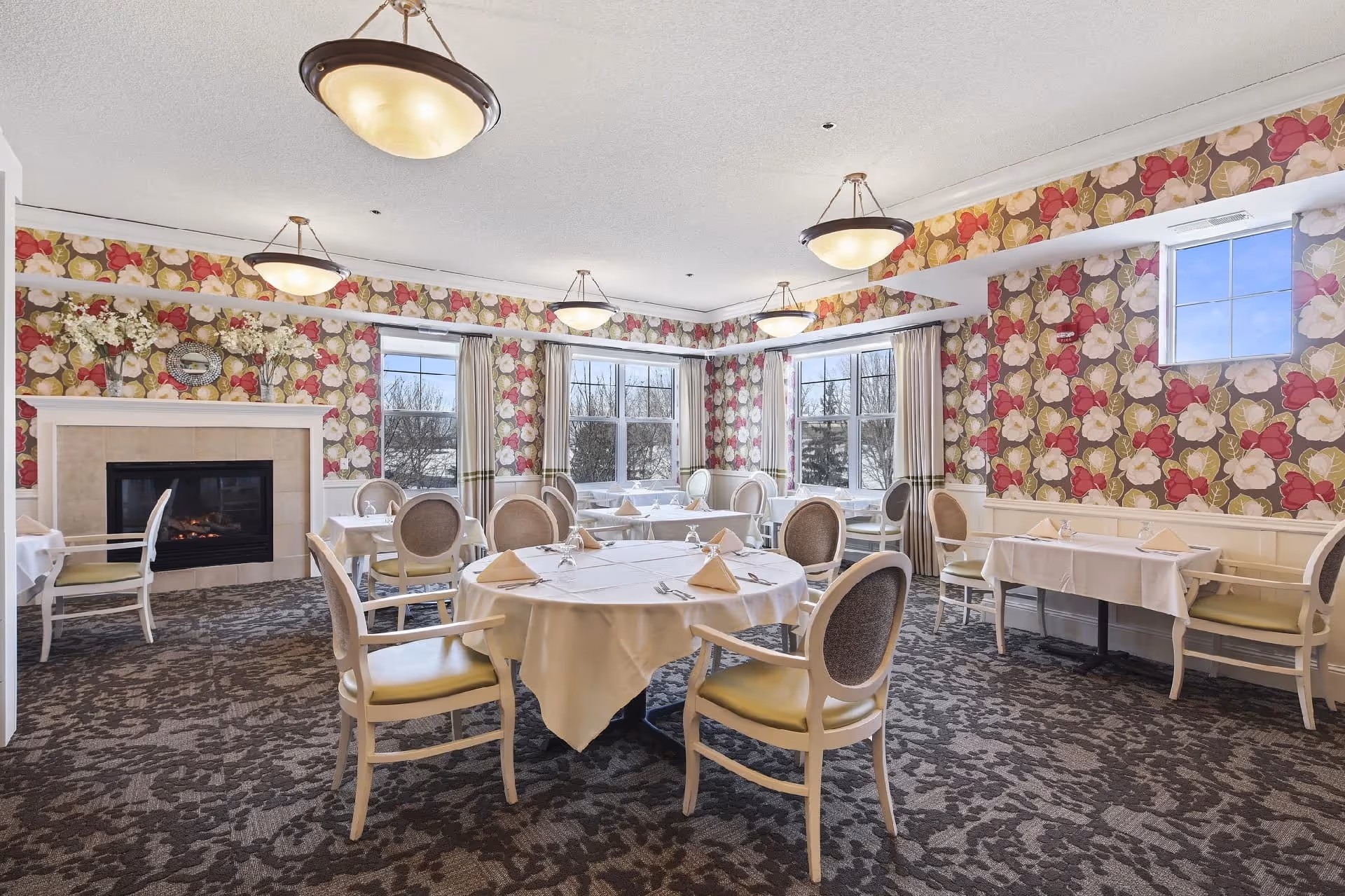 A dining room with round and rectangular tables covered with white tablecloths and set with napkins and silverware. The room features floral wallpaper with large red, pink, and beige flowers, a carpeted floor with a patterned design, several large windows letting in natural light, and a fireplace on one side. The chairs have light-colored frames and cushioned seats and backs.