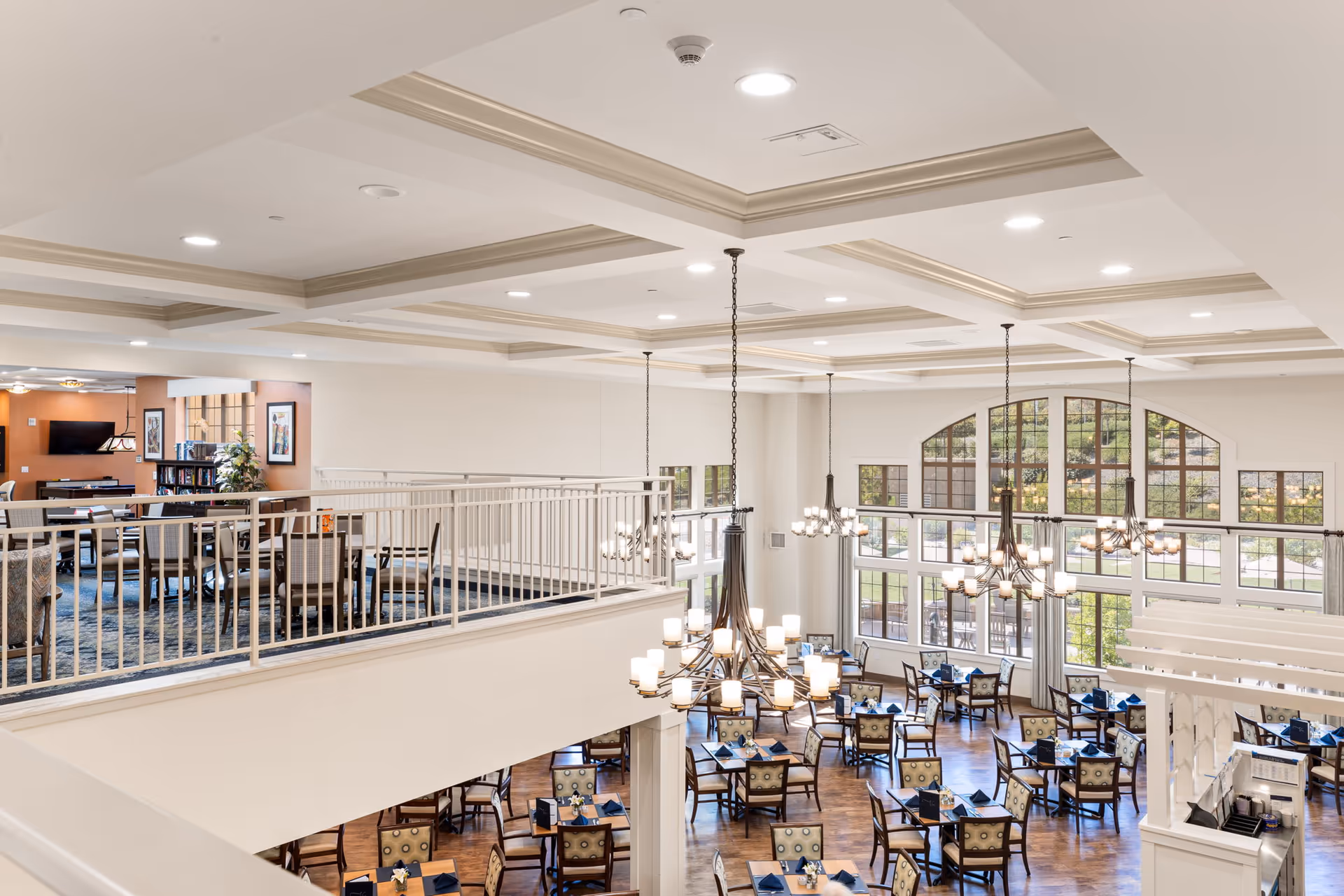 Spacious dining room in a senior living facility with multiple tables and chairs arranged neatly. Large windows with an arched design allow natural light to fill the room. Elegant chandeliers hang from the coffered ceiling. There is an upper level with additional seating and a TV area visible in the background.
